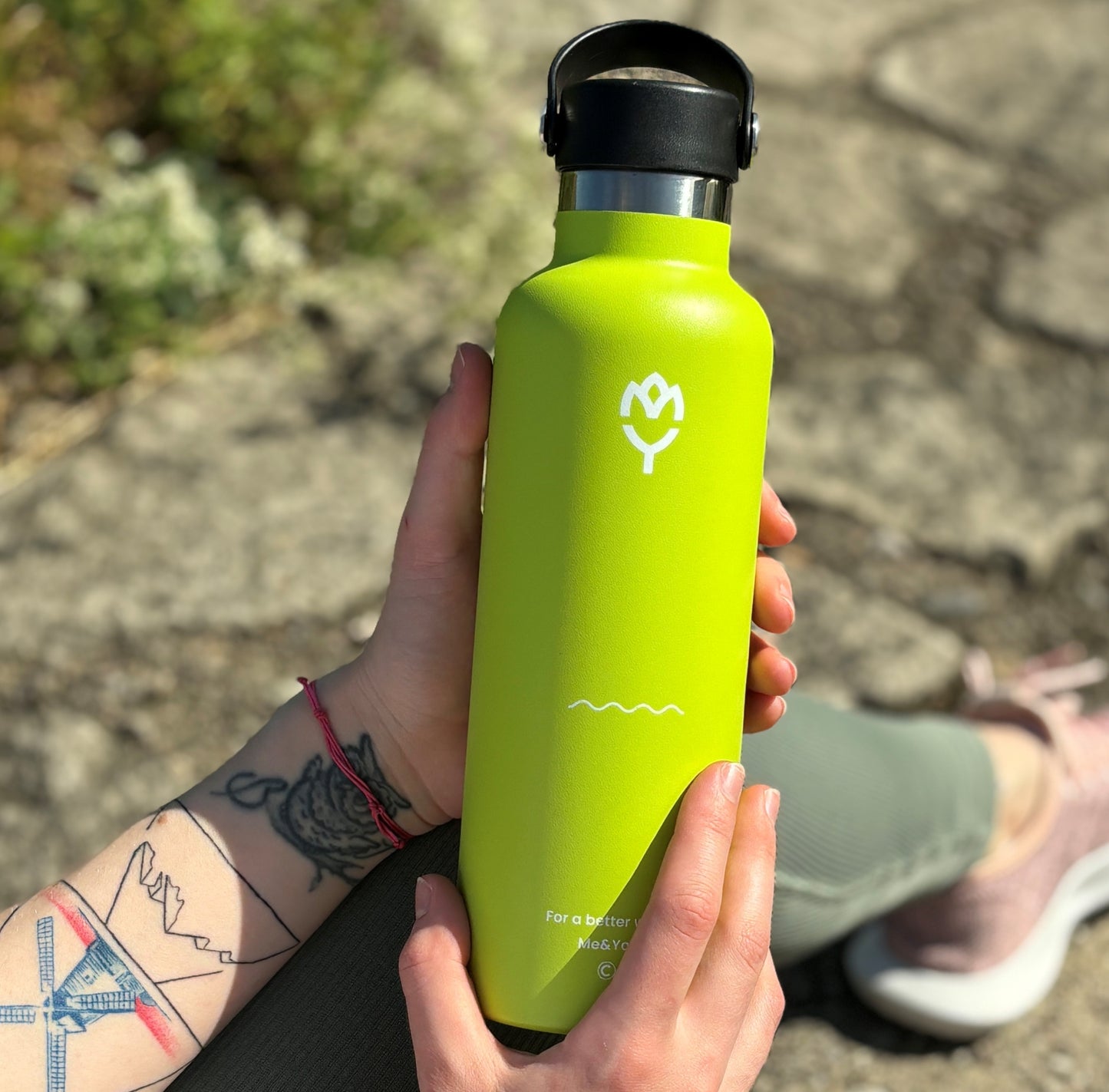Termo fľaša Iguana 750ML držaná ženskou rukou, zvýraznené telo, viečko a farba fľaše, pozadie tvorí skalnatý terén