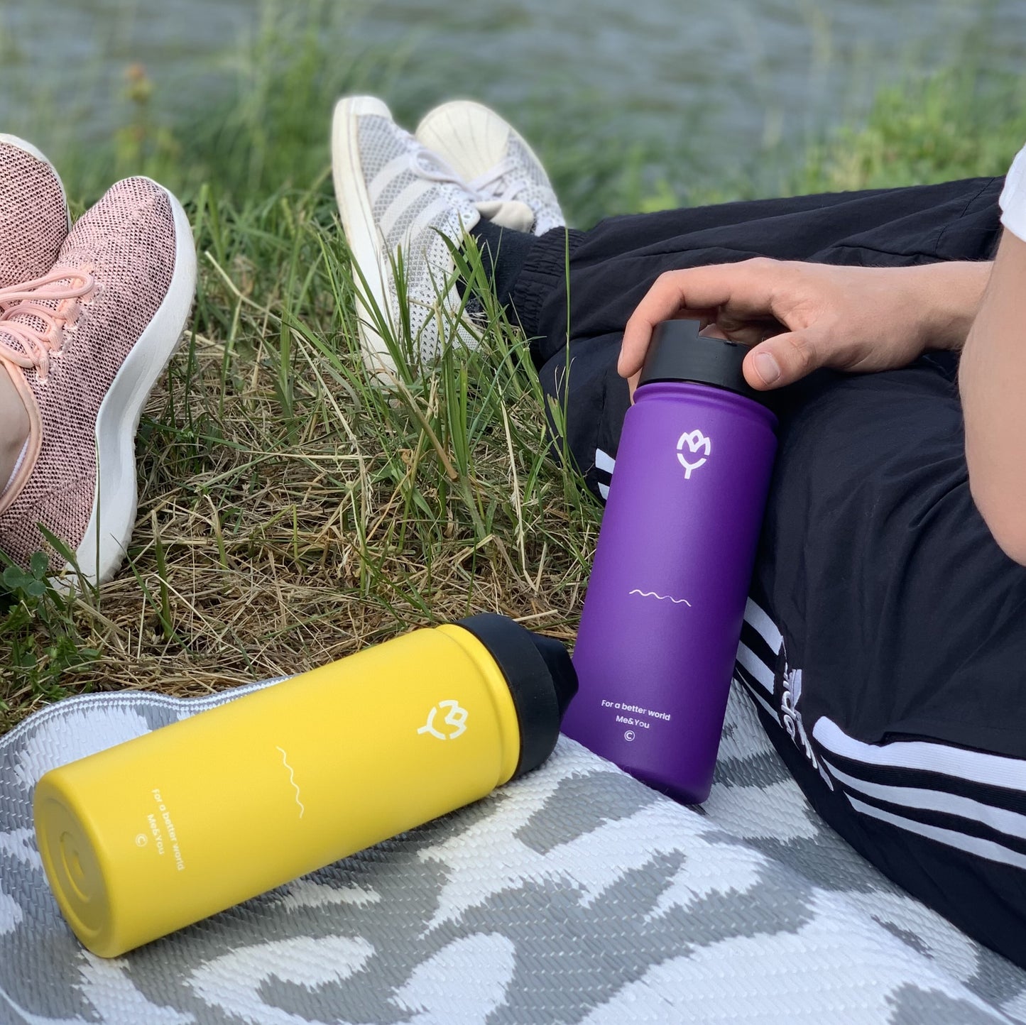 Termofľaša Gloxie 500ml (fialová) na pikniku pri rieke, viditeľné dve fľaše, nohy páru držia fľaše, ale nie sú viditeľné, zvýraznený piknikový moment
