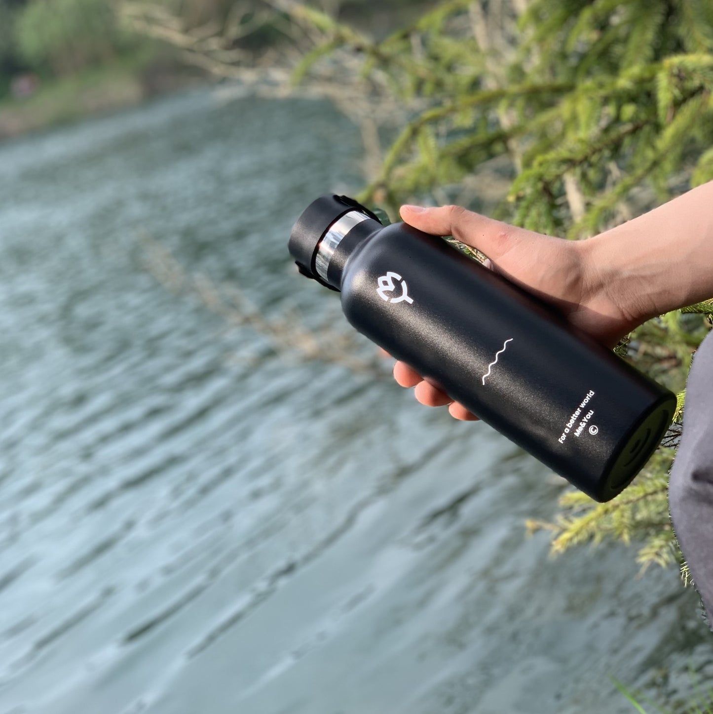 Čierna termoska Panter 750ML držaná v ruke, zvýraznená farba a telo fľaše, pozadie tvorí jazero