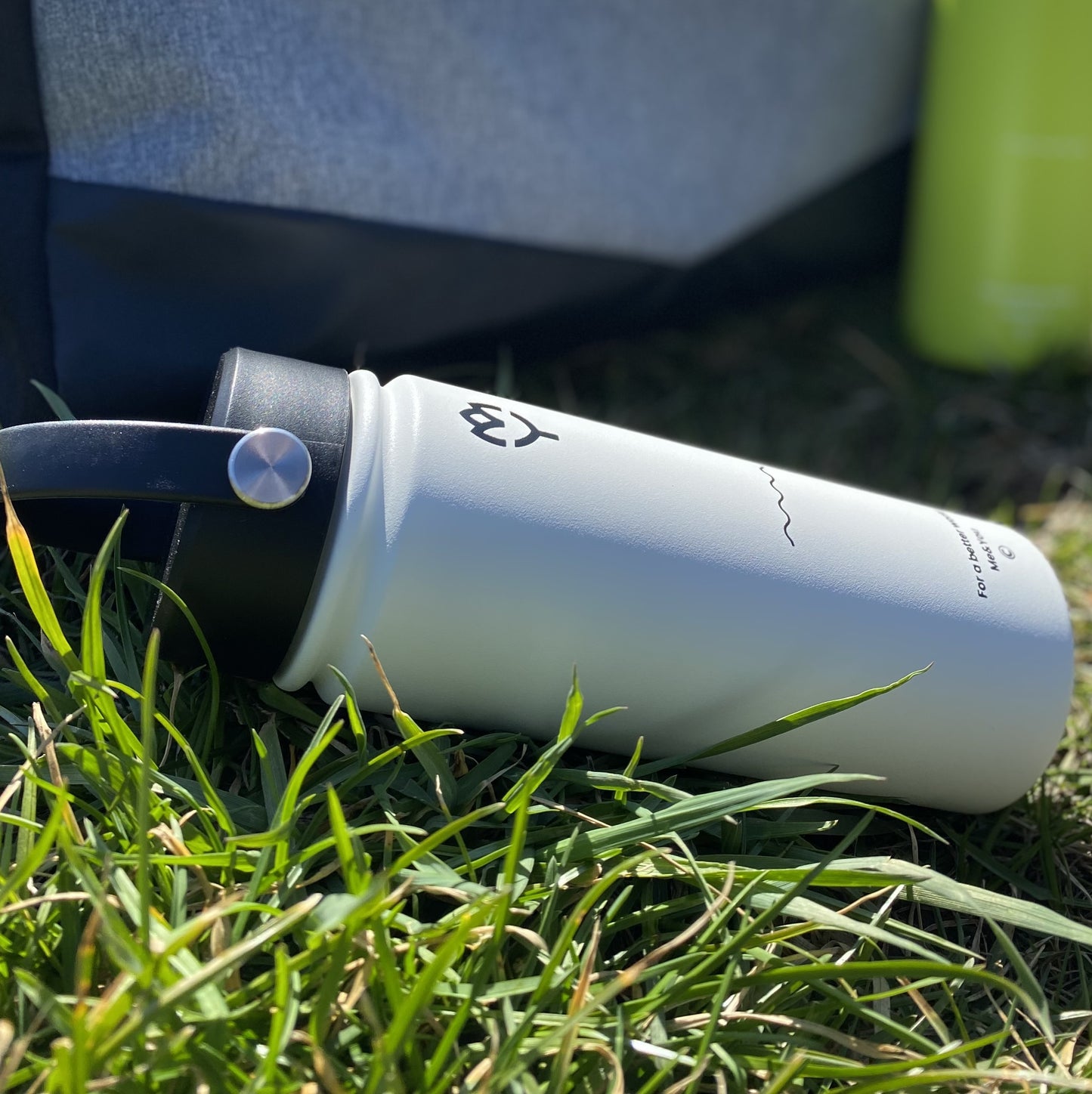 Termoska so slamkou 500ML (biela) položená v tráve, zvýraznený dizajn a farba fľaše, pozadie tvorí prírodná krajina