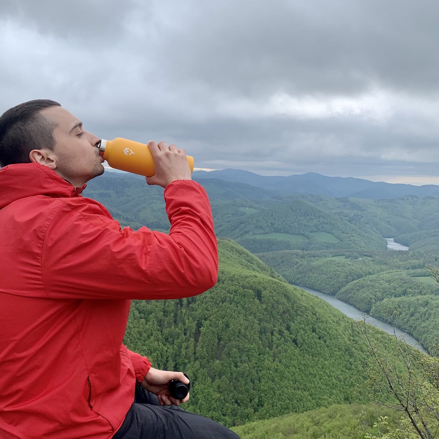 Muž na vrchole hory pije z termosky Mangifera 750ML, v pozadí horská scenéria