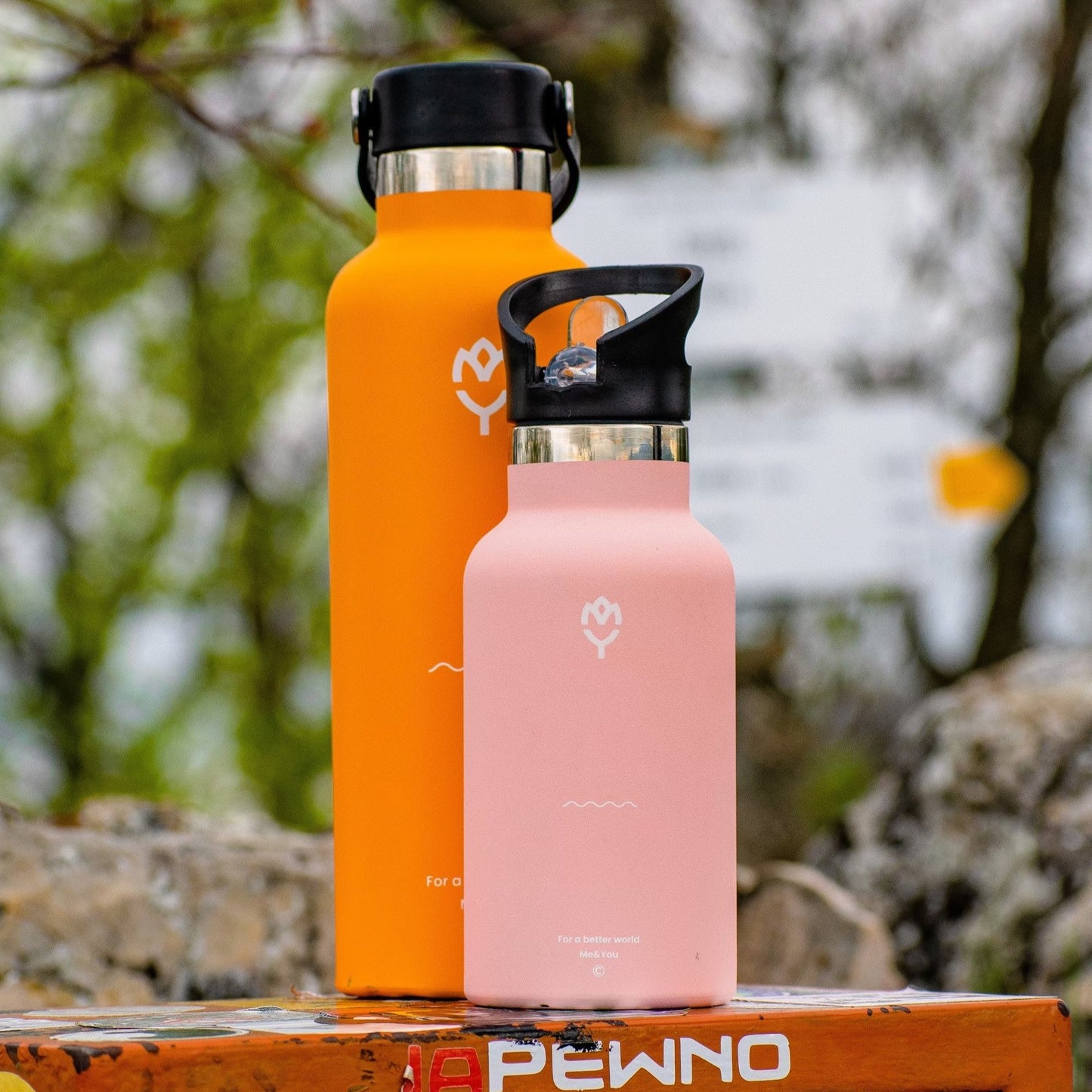 Termoska Mangifera 750ML položená vedľa ružovej fľaše na vrchole hory, bez prítomnosti ľudí, zvýraznená krása fliaš a horské prostredie