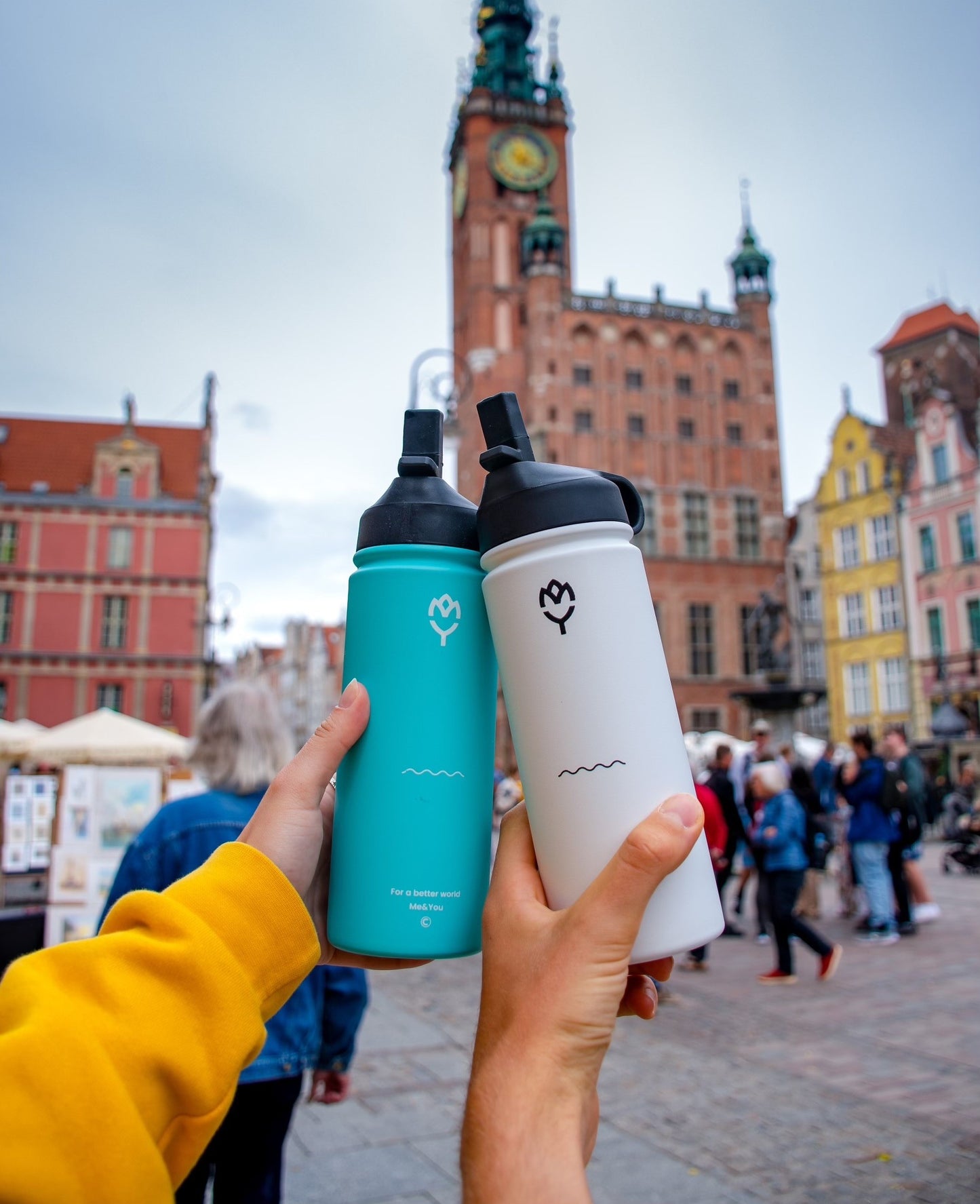 Termoska so slamkou 500ML (biela) a Adonis 500ML, držané rukami chlapca a dievčaťa, iba ruky a fľaše sú viditeľné, pozadie tvorí krásna mestská architektúra