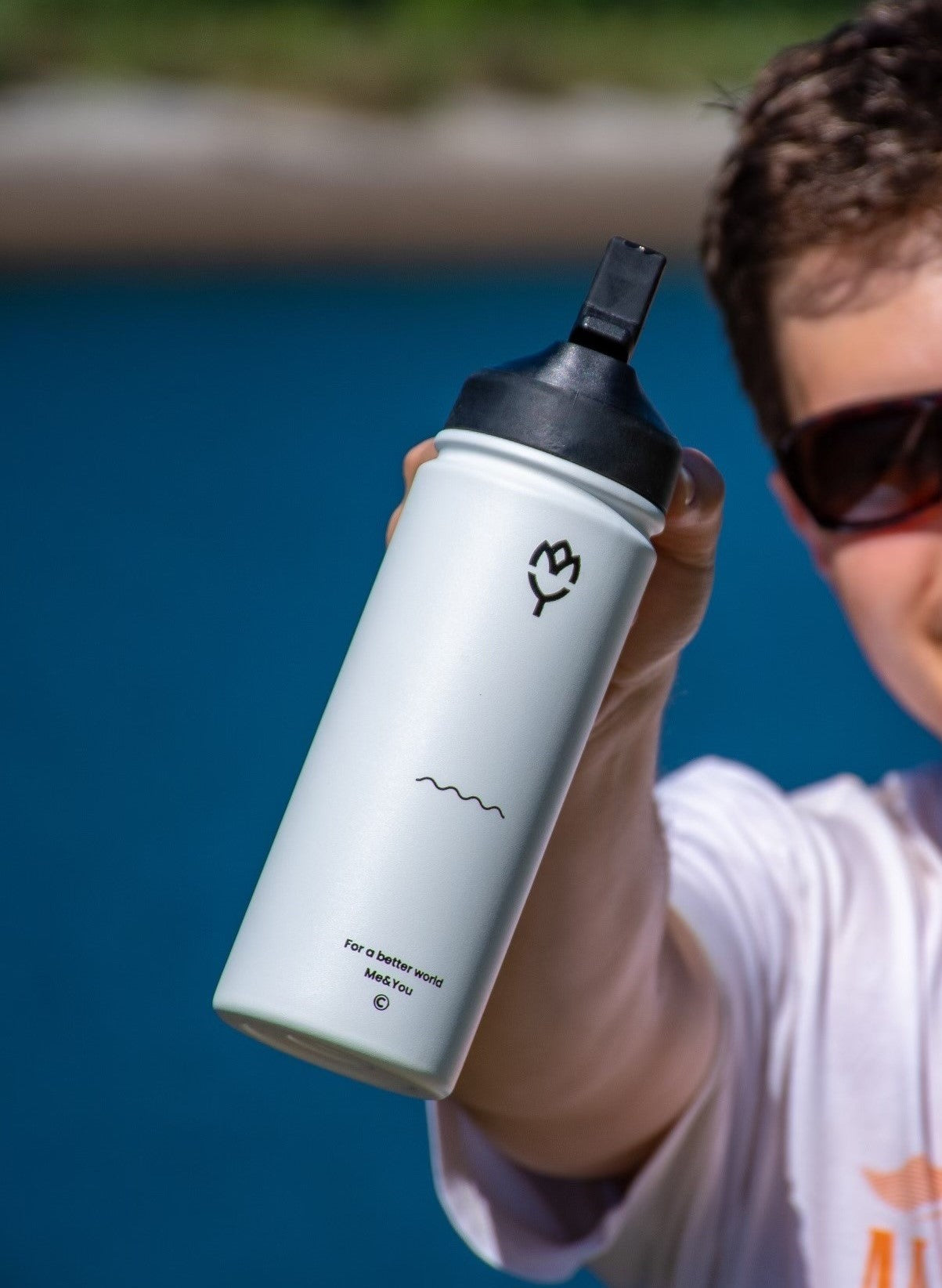Termoska so slamkou 500ML (biela) držaná mladým mužom, smerom k fotoaparátu, zvýraznená farba fľaše a spôsob držania, pozadie tvorí jazero a slnečné počasie