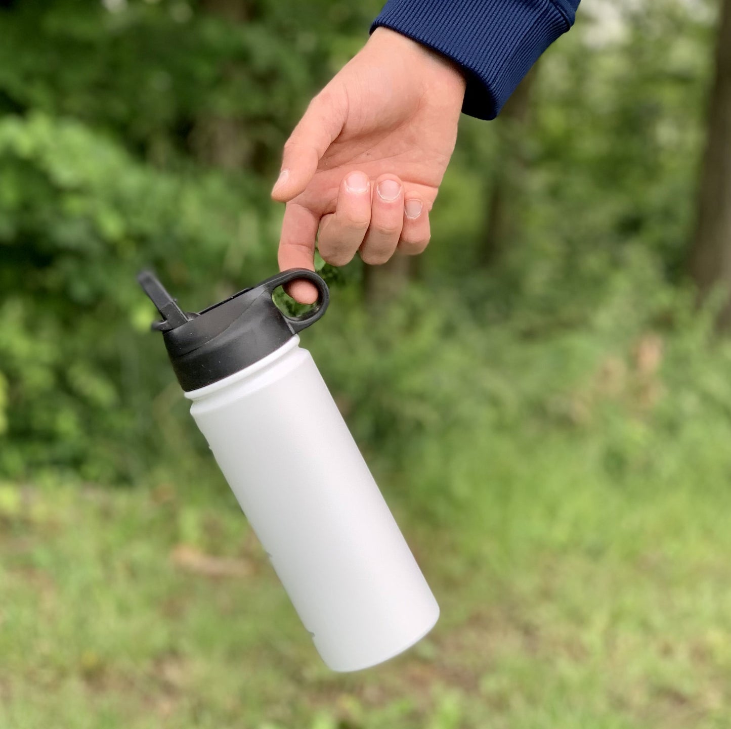 Termoska so slamkou 500ML (biela) držaná za viečko, visí vo vzduchu, zvýraznená funkčnosť viečka, pozadie tvorí les