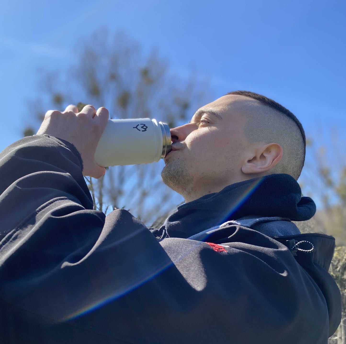Termoska so slamkou 500ML (biela) mladým mužom pité, pozadie tvorí časť stromu a obloha, snímané zospodu