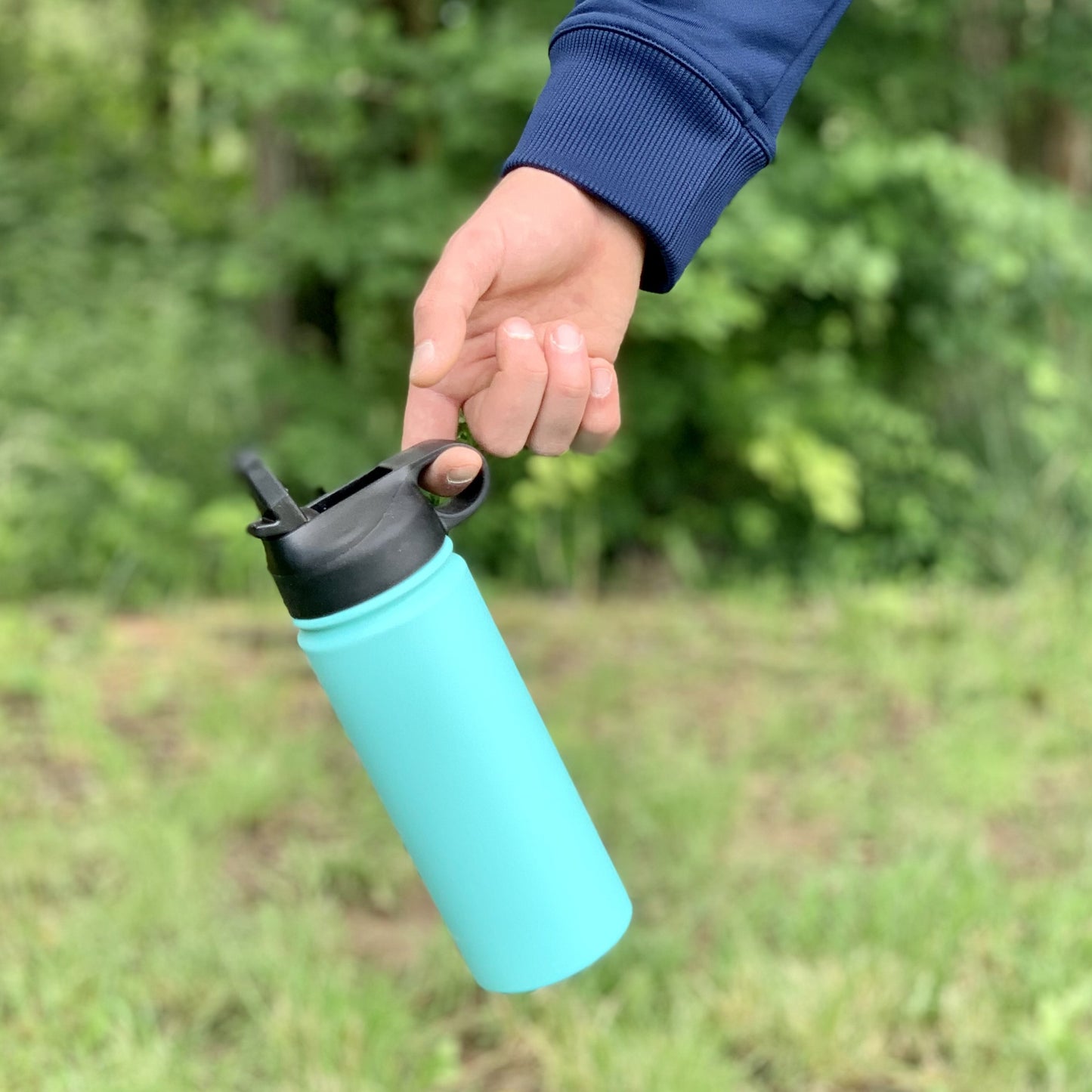 Detská termoska so slamkou 500ML (modrá) držaná za viečko, visí vo vzduchu, pozadie tvorí les, zvýraznená funkčnosť viečka
