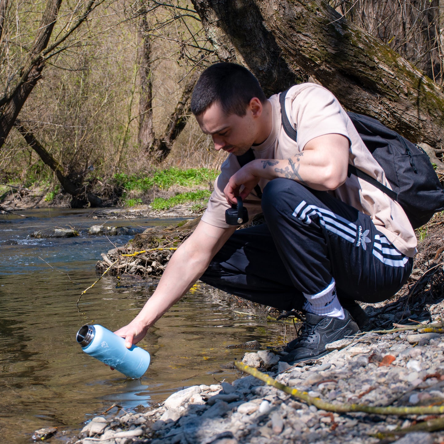 Muž s ruksakom pri rieke sa nakláňa k vode, aby naplnil modrú termosku Bluo – zvýraznená funkčnosť, farba a outdoor použitie