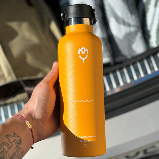 Termoska Mangifera 750ML držaná v ruke v slnečnom počasí, zvýraznené telo, farba a viečko fľaše, pozadie tvorí batožinový priestor auta