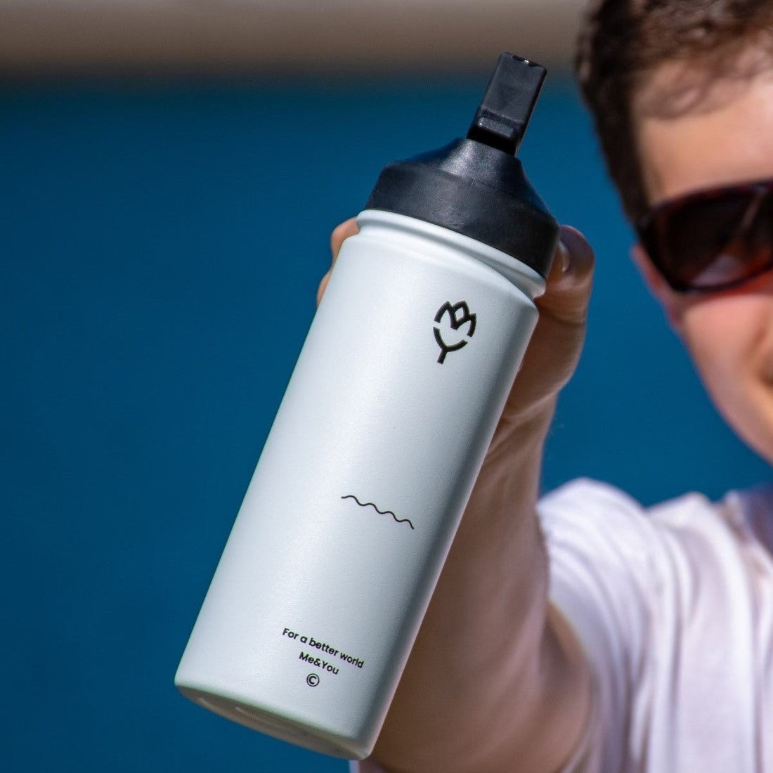  Termoska so slamkou 500ML (biela) držaná mladým mužom, smerom k fotoaparátu, zvýraznená farba fľaše a spôsob držania, pozadie tvorí jazero a slnečné počasie