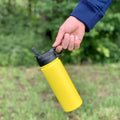 Termoska pre deti 500ml (Slnecnica) držaná za viečko, visí vo vzduchu, pozadie tvorí les, zvýraznený tvar a veľkosť fľaše

