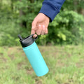 Detská termoska so slamkou 500ML (modrá) držaná za viečko, visí vo vzduchu, pozadie tvorí les, zvýraznená funkčnosť viečka

