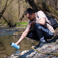 Muž s ruksakom pri rieke sa nakláňa k vode, aby naplnil modrú termosku Bluo – zvýraznená funkčnosť, farba a outdoor použitie
